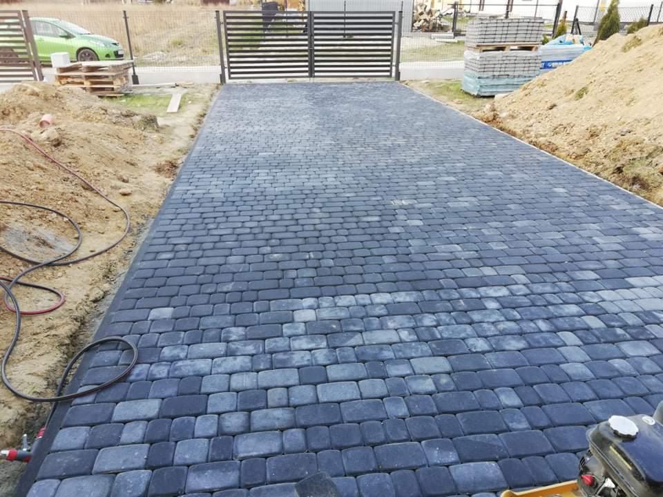 Ułożona, szara kostka brukowa prowadząca do bramy w Lubaczowie, widoczna ubijarka wibracyjna i palety kostki w tle.