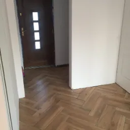 Ułożona w jodełkę, drewnopodobna podłoga z płytek, widok z progu na brązowe drzwi z trzema okienkami i białe ściany w trakcie prac wykończeniowych.