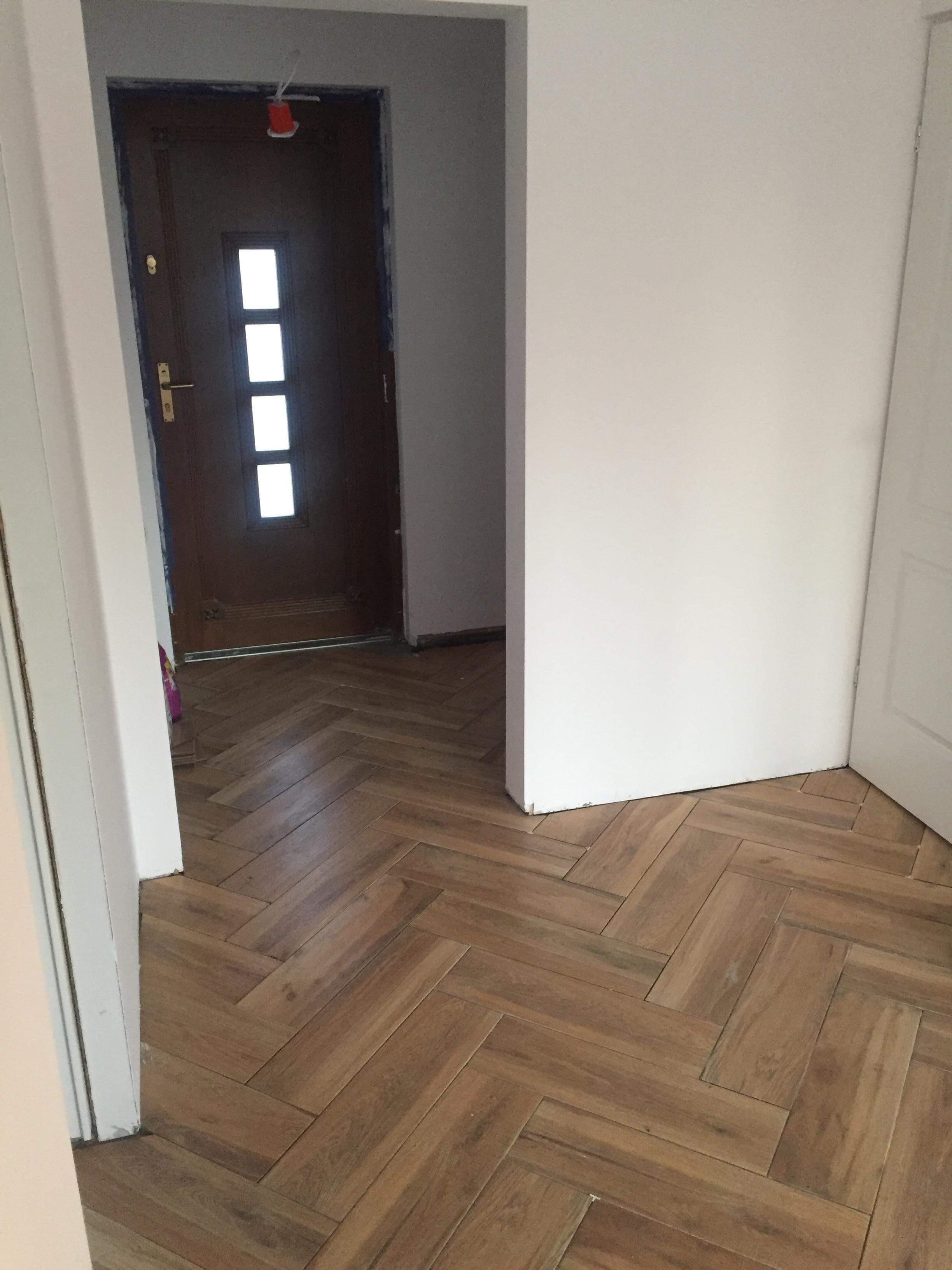 Ułożona w jodełkę, drewnopodobna podłoga z płytek, widok z progu na brązowe drzwi z trzema okienkami i białe ściany w trakcie prac wykończeniowych.