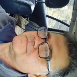 Mężczyzna w okularach siedzi w samochodzie, ubrany w szarą bluzę i niebieską koszulkę. Widoczna część twarzy, włosy i wnętrze pojazdu z kamiennym murem w tle.