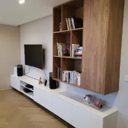 Nowoczesna zabudowa ściany z białymi szafkami dolnymi i drewnianym regałem wiszącym, telewizor na ścianie, parkiet w jodełkę na podłodze.