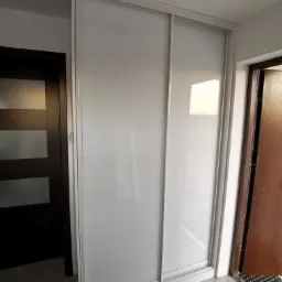 Biała szafa przesuwna z matowego szkła, wkomponowana w wąskie pomieszczenie między dwiema parami drzwi.