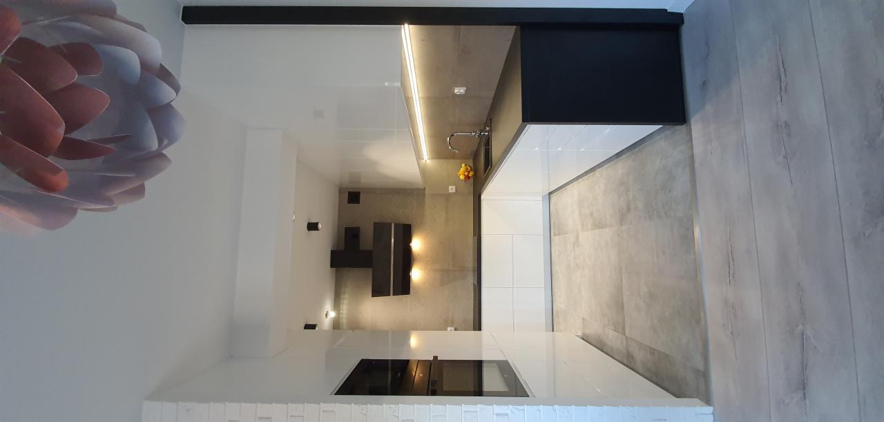 Nowoczesna kuchnia z białymi szafkami i blatem, szarą ścianą oraz drewnianą podłogą. W tle widoczny czarny okap i oświetlenie punktowe.