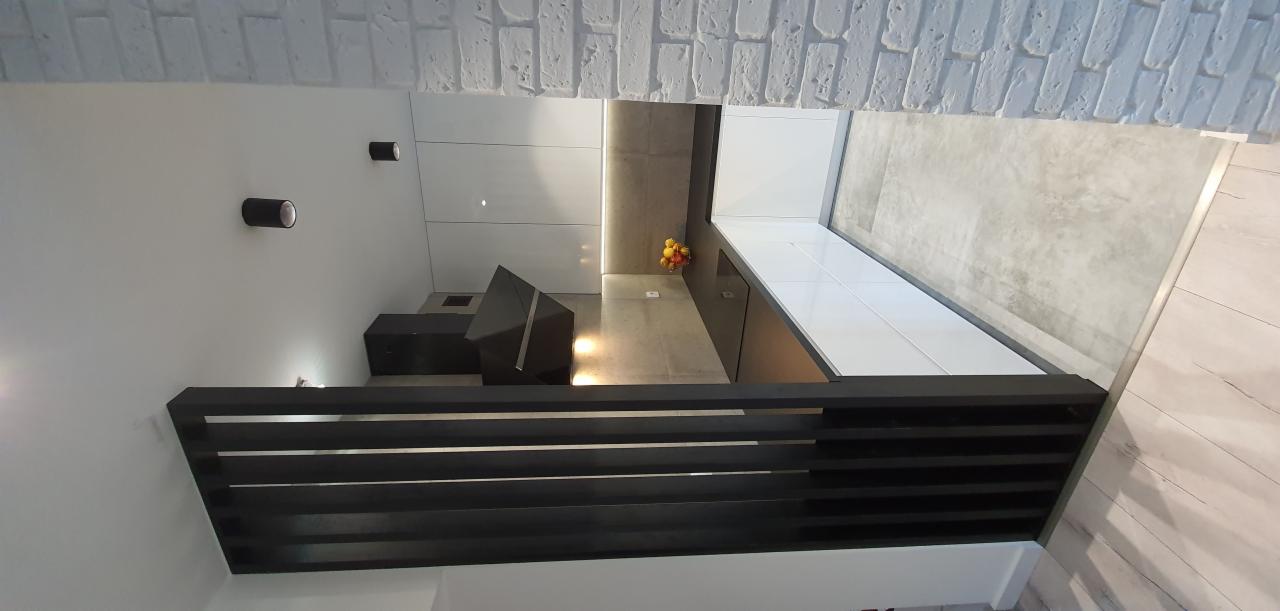 Nowoczesna kuchnia z białymi frontami szafek, szarym blatem i betonową ścianą, oświetlona punktowymi lampami.