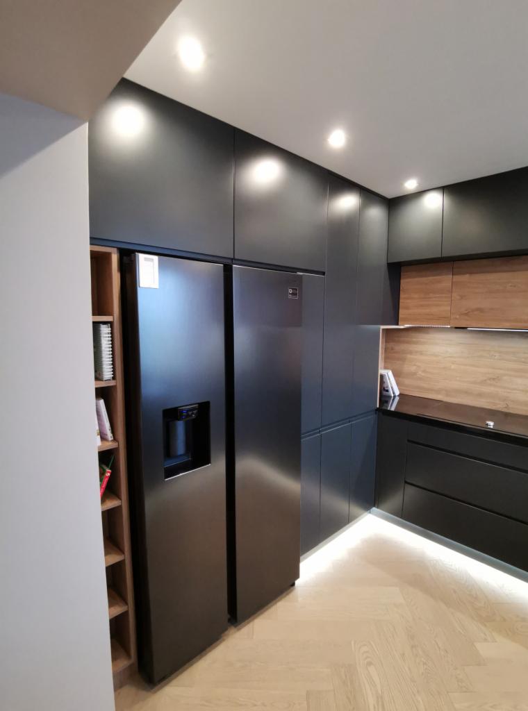 Nowoczesna, czarna kuchnia z lodówką side-by-side i drewnianymi akcentami, oświetleniem LED pod szafkami i parkietem w jodełkę.