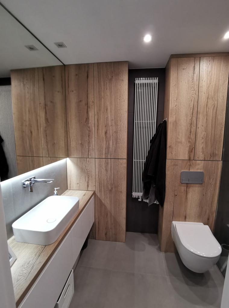 Nowoczesna łazienka z drewnianymi panelami ściennymi, białą umywalką nablatową i podwieszaną toaletą, oświetlenie LED pod lustrem.