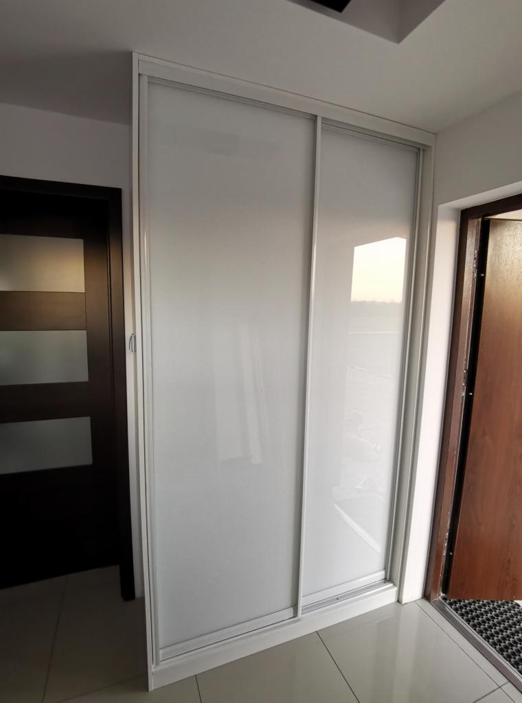 Biała szafa przesuwna z matowego szkła, wkomponowana w wąskie pomieszczenie między dwiema parami drzwi.