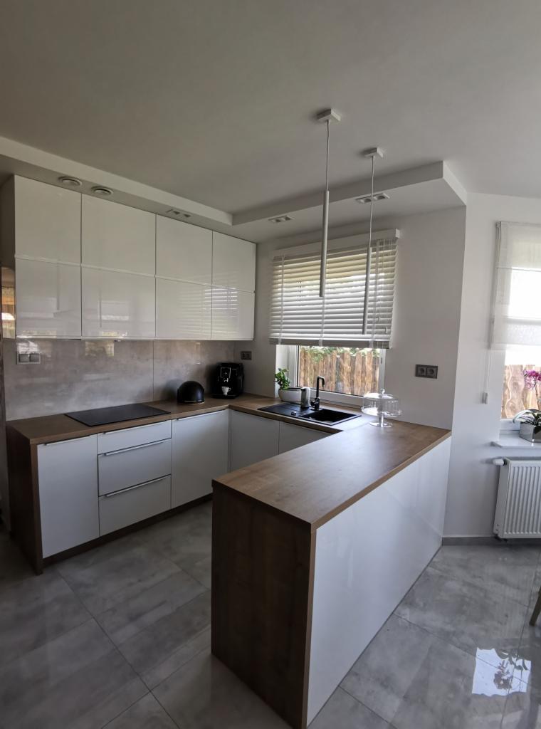 Nowoczesna kuchnia z białymi, błyszczącymi frontami szafek i drewnianym blatem, widoczna czarna armatura i ekspres do kawy.