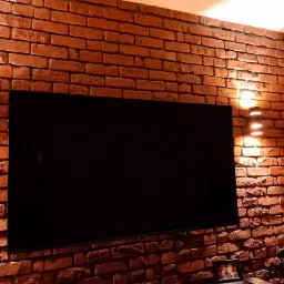 Telewizor zawieszony na ścianie z cegły, oświetlonej przez dwa kinkiety, tworzące ciepłą atmosferę w pomieszczeniu.