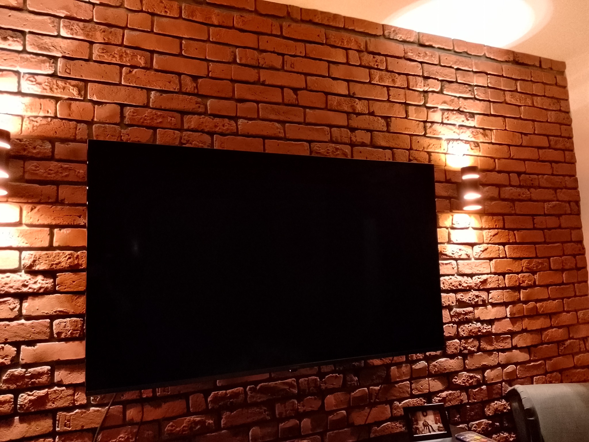Telewizor zawieszony na ścianie z cegły, oświetlonej przez dwa kinkiety, tworzące ciepłą atmosferę w pomieszczeniu.