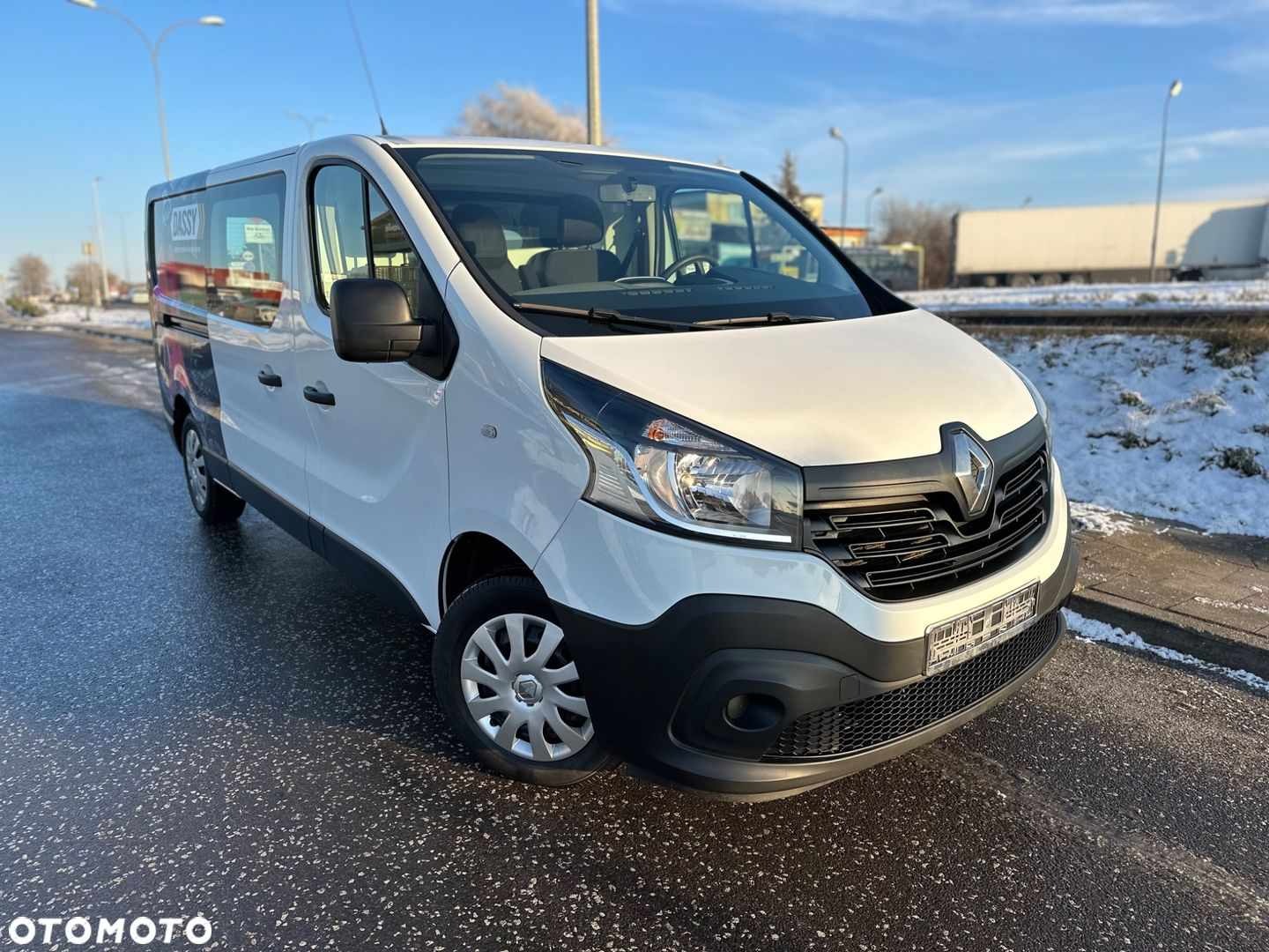Biały bus Renault Trafic z czarnymi elementami, zaparkowany na mokrym asfalcie w zimowy dzień, widoczny z przodu pod lekkim kątem, z napisem 'DASSY' na boku.