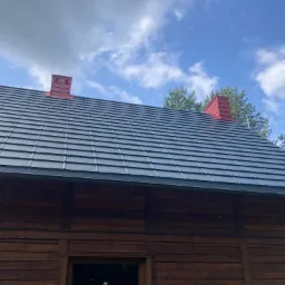 Ciemnoszary dach z blachodachówki na drewnianym domu, z czerwonymi kominami, na tle błękitnego nieba z chmurami i zieleni drzew.