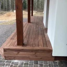 Zadaszony taras drewniany z brązowymi słupami w Atenach, widok z poziomu kostki brukowej, w tle las.