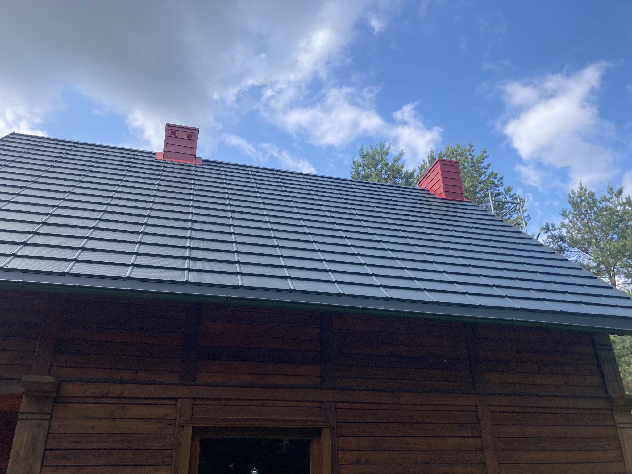 Ciemnoszary dach z blachodachówki na drewnianym domu, z czerwonymi kominami, na tle błękitnego nieba z chmurami i zieleni drzew.