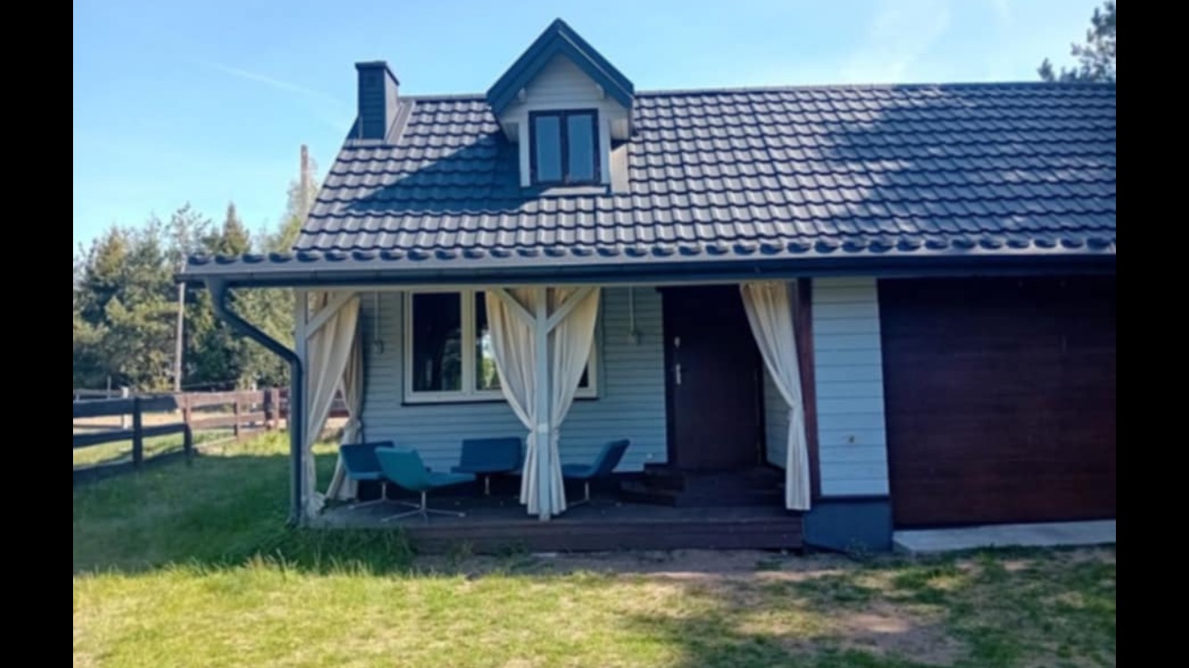 Dom z ciemną dachówką i jasnymi zasłonami na tarasie. Widoczne niebieskie krzesła i zadbany trawnik. Ujęcie w słoneczny dzień, z perspektywy trawnika.