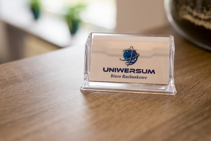 Wizytownik z wizytówkami firmy 'Uniwersum Biuro Rachunkowe' z logo przedstawiającym globus, na drewnianym biurku w jasnym otoczeniu.