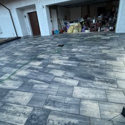 M-K Kompleksowe usługi brukarskie - Ułożona, szara kostka brukowa na podjeździe przed domem z otwartym garażem. Widoczny fragment trawnika i osoba w odblaskowej kamizelce. W tle dom z czerwonym dachem.