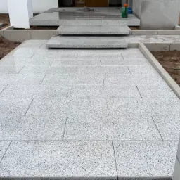 Świeżo ułożona, jasna kostka granitowa prowadząca do schodów wejściowych; widoczne słupki ogrodzeniowe z betonu architektonicznego po bokach ścieżki.