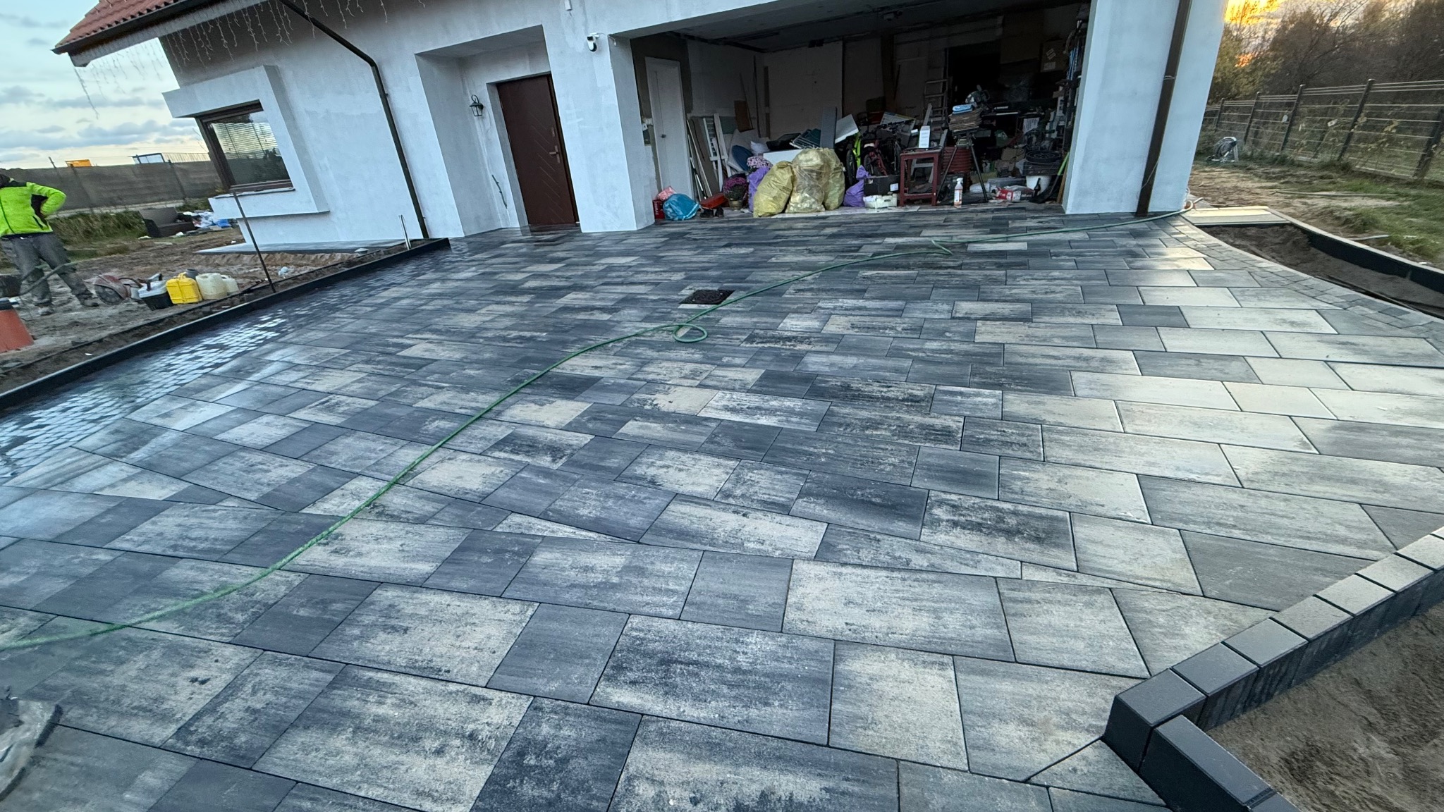 Ułożona, szara kostka brukowa na podjeździe przed domem z otwartym garażem. Widoczny fragment trawnika i osoba w odblaskowej kamizelce. W tle dom z czerwonym dachem.