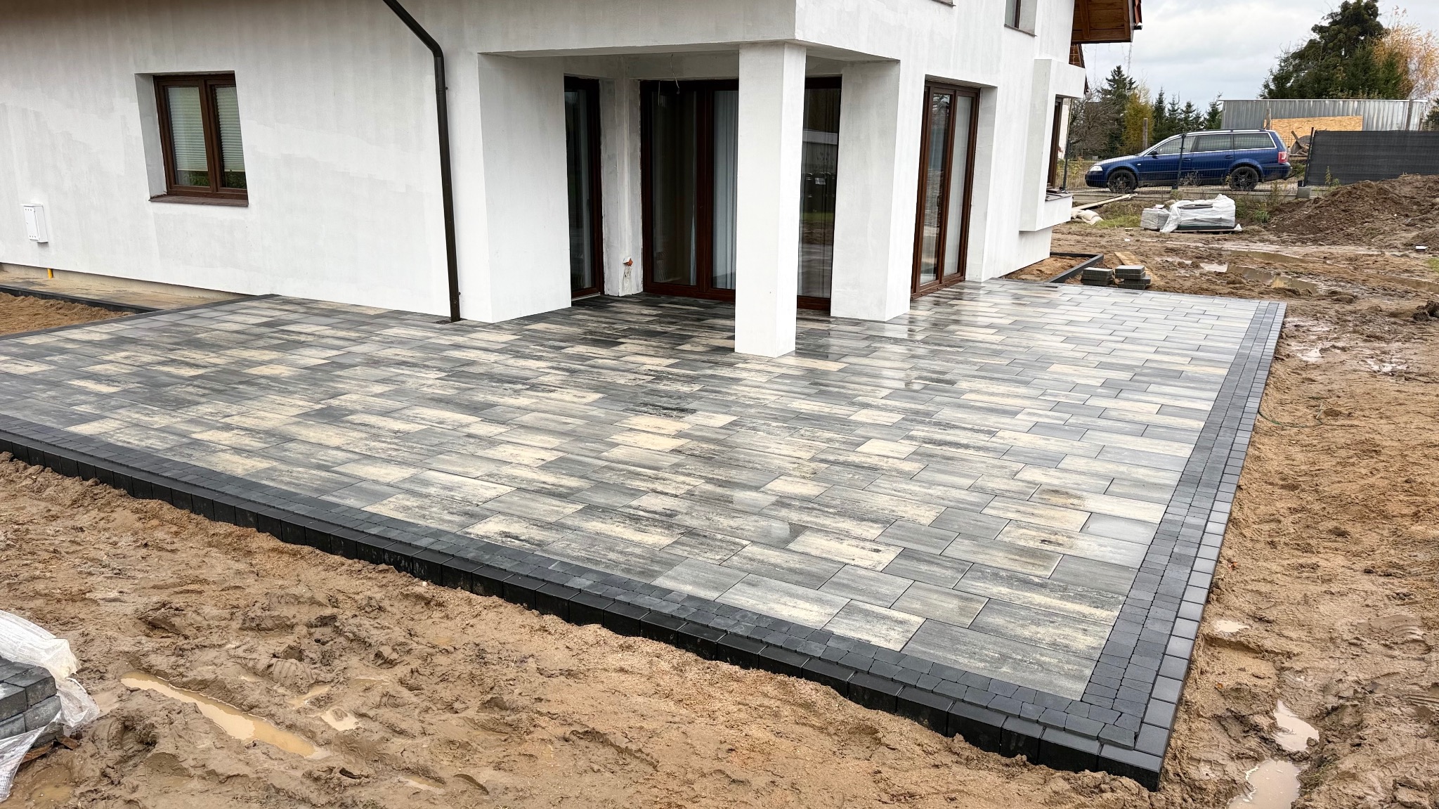 Nowoczesny taras z kostki brukowej w odcieniach szarości, obramowany ciemną kostką. Widok na biały dom z brązowymi oknami i drzwi.