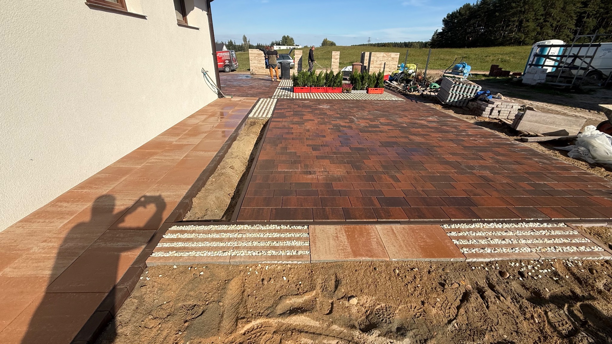 Częściowo ułożona, brązowa kostka brukowa na posesji. Widoczne obrzeża, podsypka z kamyków i fragmenty wykopu. W tle budynek i osoby przy pracach wykończeniowych.