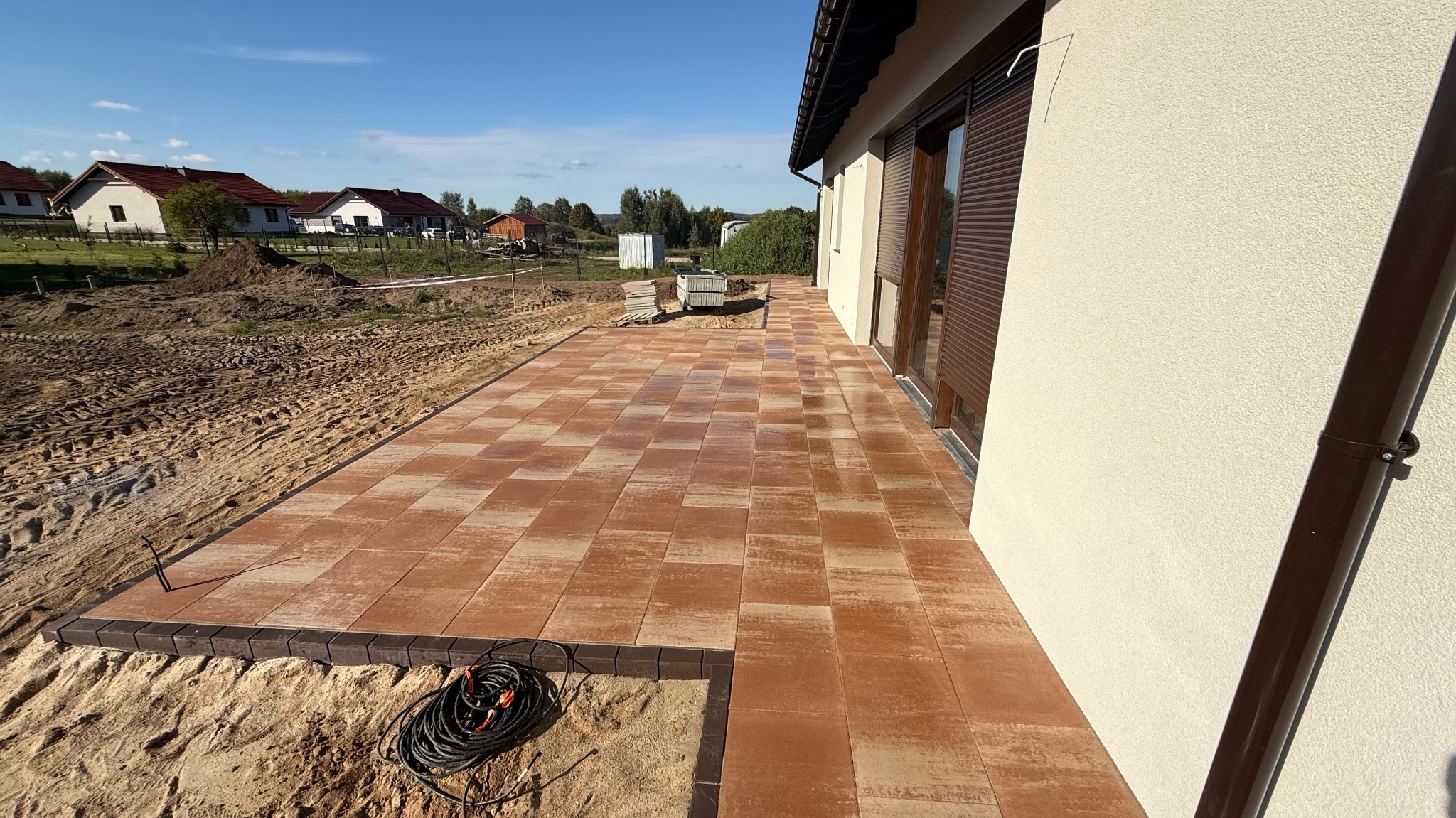 Ułożona, brązowa kostka brukowa tworząca ścieżkę przy budynku z brązowymi roletami. W tle widoczny teren przygotowany pod dalsze prace.