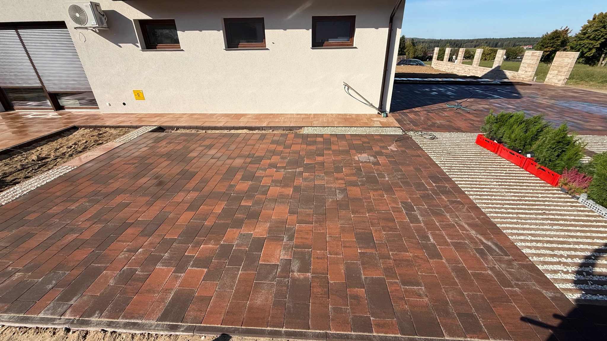 Częściowo ułożona, brązowo-czerwona kostka brukowa na posesji. Widoczne przygotowane podłoże i obrzeża. W tle budynek z klimatyzatorem i fragment ogrodzenia.