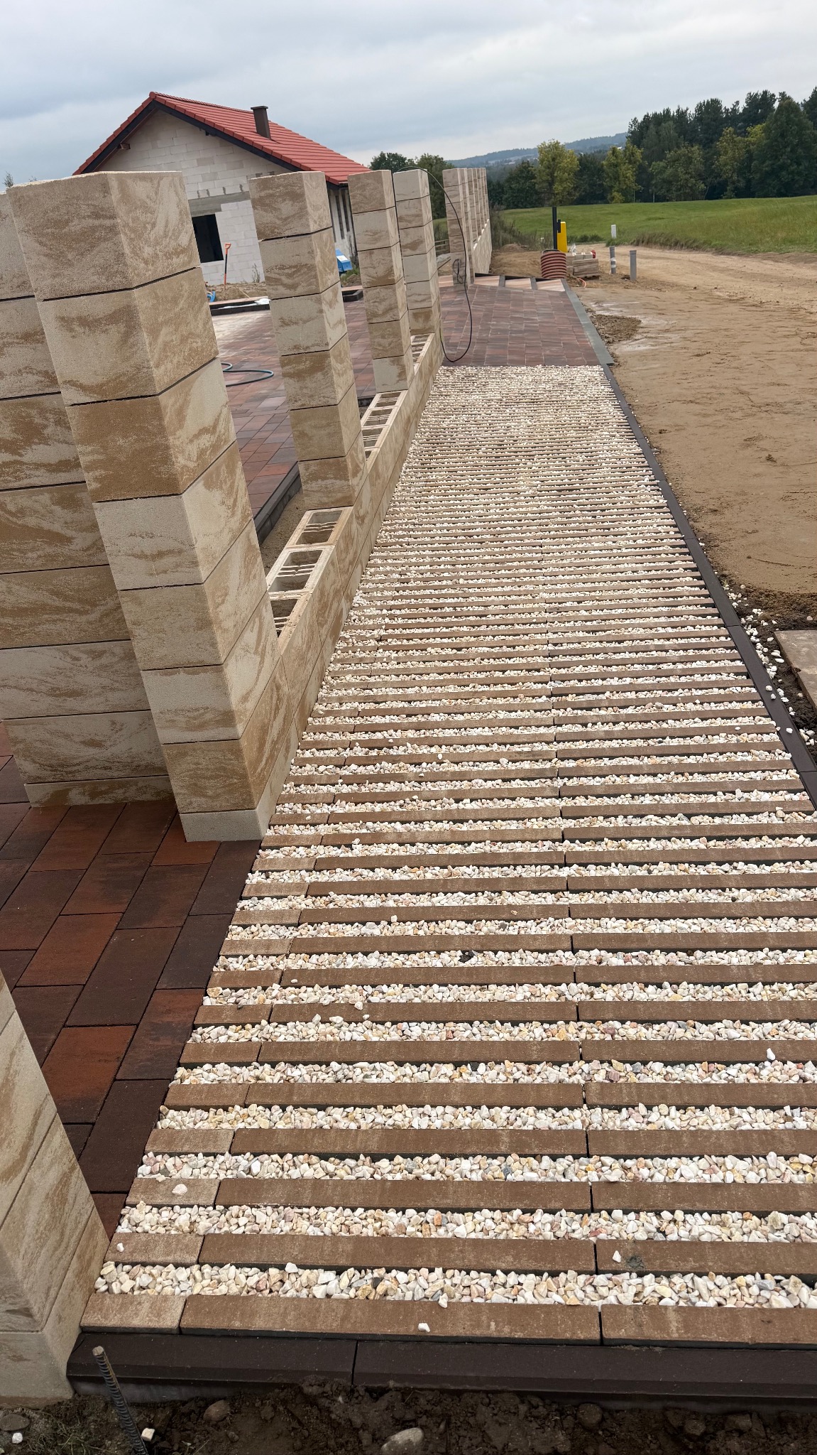 Nowoczesna aranżacja ścieżki z kostki betonowej z białym żwirem, prowadząca wzdłuż ogrodzenia z pustaków o piaskowej barwie. Widok na dom w tle.