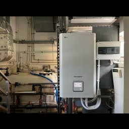 Bat-Pol - Biała pompa ciepła LG Therma V zamontowana w pomieszczeniu technicznym, widoczne rury, zawory i zbiornik buforowy.