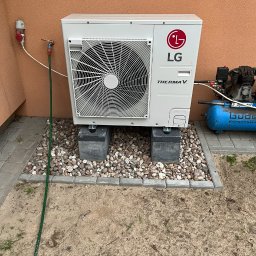 Bat-Pol - Zewnętrzna jednostka pompy ciepła LG Therma V, umieszczona na betonowych bloczkach i otoczona kamieniami, z widocznym podłączeniem do instalacji i kompresorem powietrza w tle.