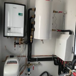 Bat-Pol - Zestaw urządzeń grzewczych: biały bojler Sunsystem, kocioł Vaillant z zieloną naklejką i filtr do wody, połączone czarnymi rurami w pomieszczeniu.