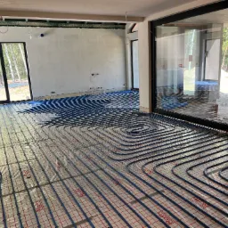 Rozłożone, niebieskie rury ogrzewania podłogowego na folii z czerwoną kratką w surowym wnętrzu domu z dużymi oknami tarasowymi.