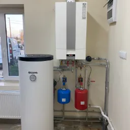 Biały kocioł gazowy Wolf z szarym pasem na ścianie, obok biały zasobnik Kospel z czarnymi elementami, czerwony i niebieski zbiornik wyrównawczy, instalacja rurowa, okno z widokiem na samochód...