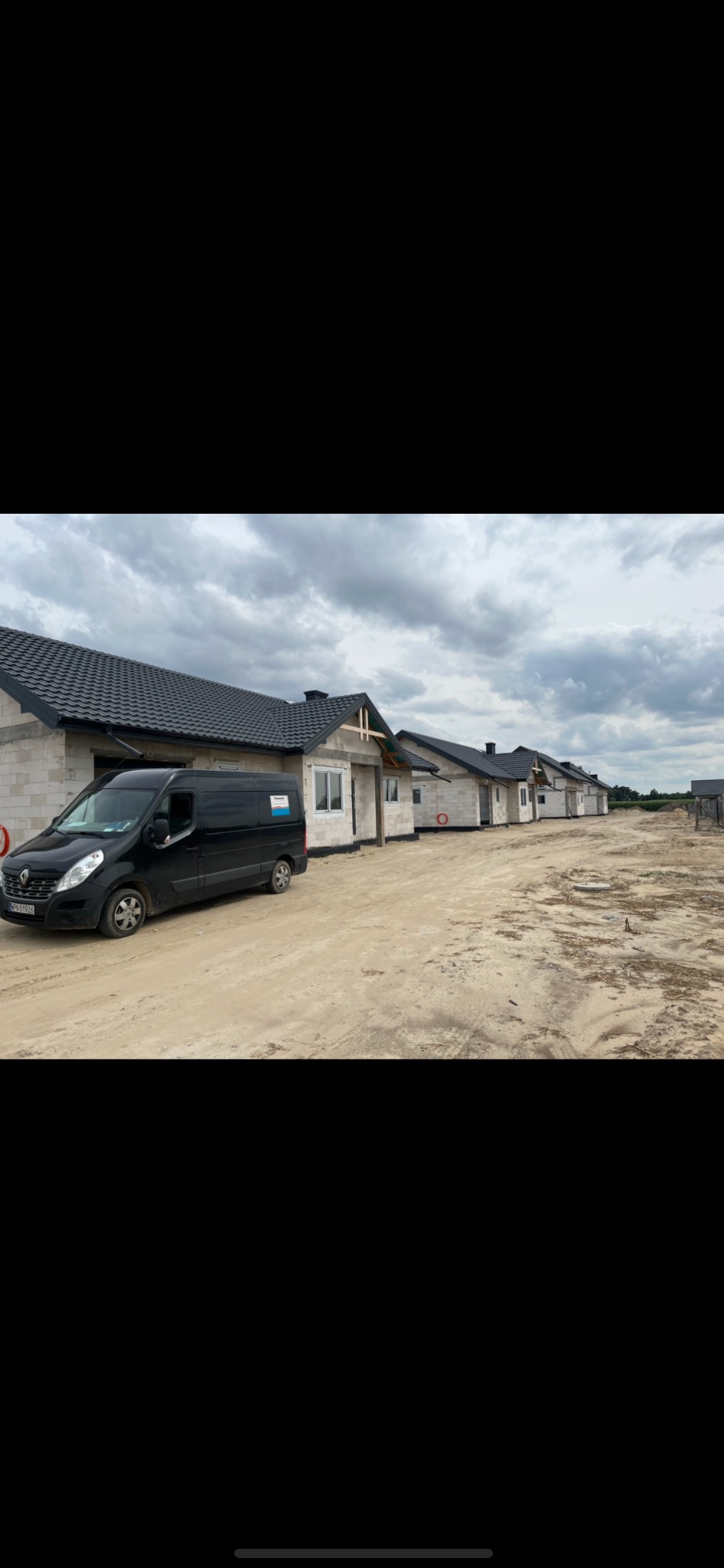 Rząd powstających domów jednorodzinnych z czarnymi dachami, z zaparkowanym czarnym vanem z naklejką firmy na boku, na piaszczystym terenie budowy pod pochmurnym niebem.