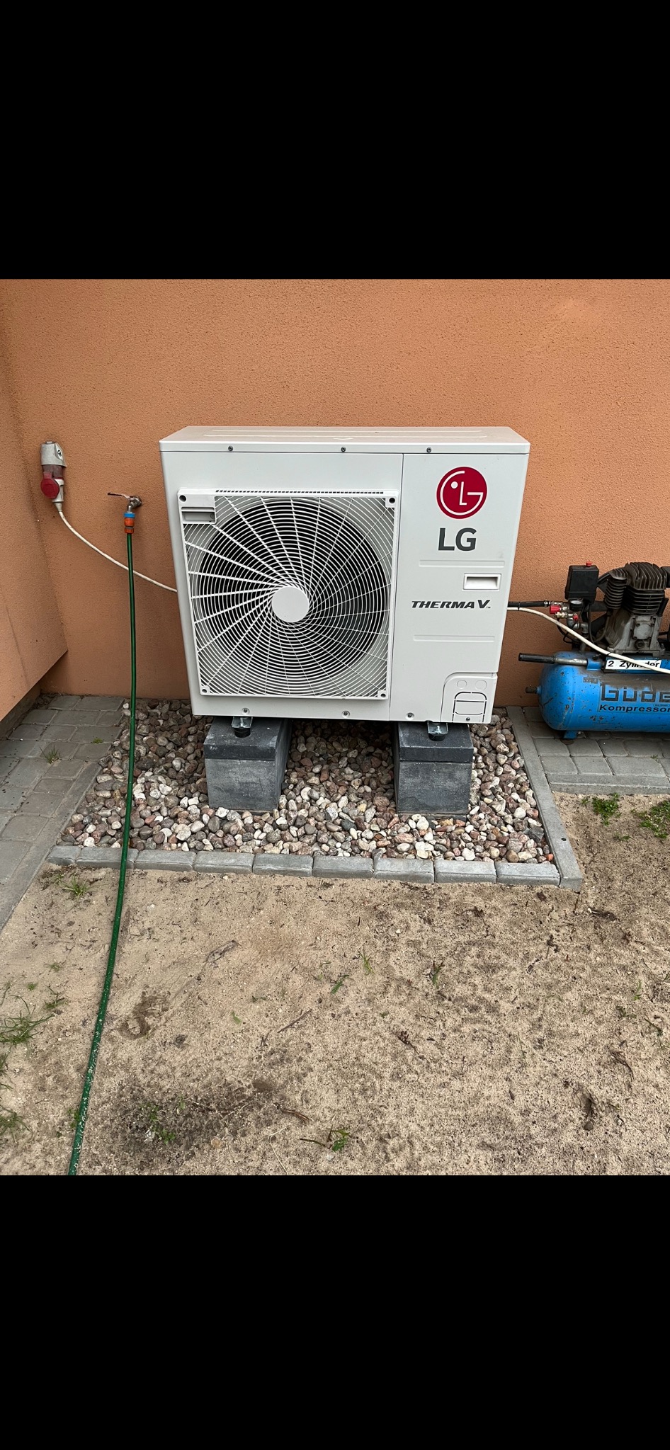 Zewnętrzna jednostka pompy ciepła LG Therma V, umieszczona na betonowych bloczkach i otoczona kamieniami, z widocznym podłączeniem do instalacji i kompresorem powietrza w tle.