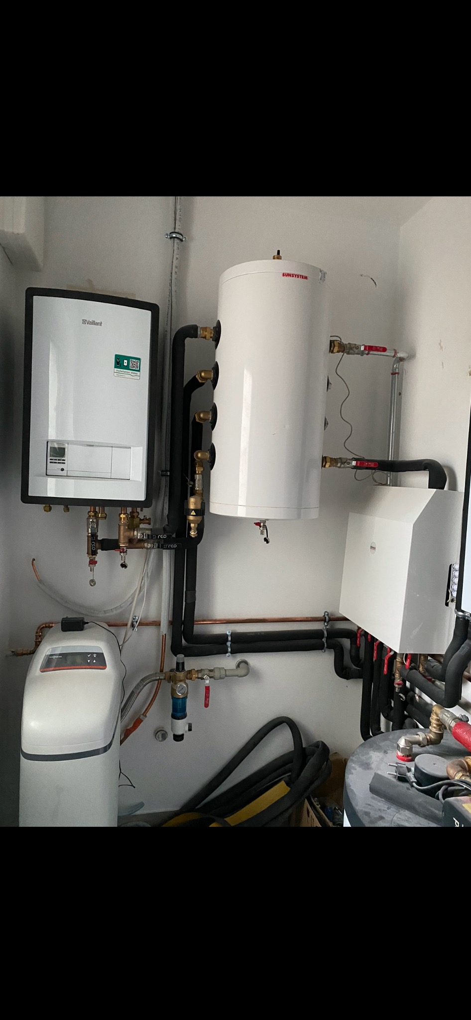 Zestaw urządzeń grzewczych: biały bojler Sunsystem, kocioł Vaillant z zieloną naklejką i filtr do wody, połączone czarnymi rurami w pomieszczeniu.