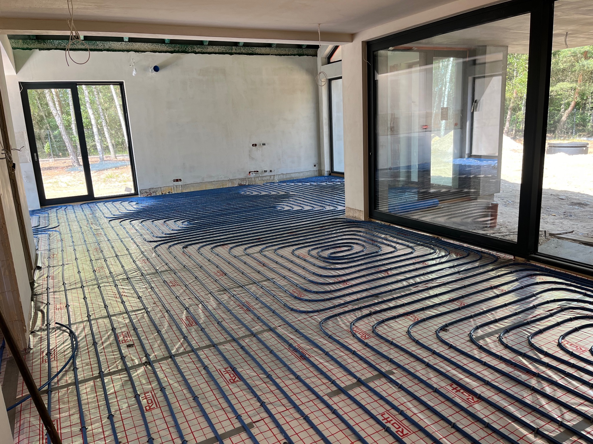 Rozłożone, niebieskie rury ogrzewania podłogowego na folii z czerwoną kratką w surowym wnętrzu domu z dużymi oknami tarasowymi.