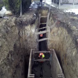 Głęboki wykop zabezpieczony szalunkami stalowymi, dwóch pracowników w kaskach i kombinezonach wewnątrz, na górze osoba nadzorująca z niwelatorem, częściowo zasypany śniegiem teren.
