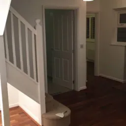 Wnętrze korytarza po malowaniu: białe ściany, drewniana podłoga, schody z białą balustradą, otwarte drzwi do kolejnych pomieszczeń, widoczny pędzel na schodach.