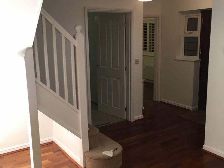 Wnętrze korytarza po malowaniu: białe ściany, drewniana podłoga, schody z białą balustradą, otwarte drzwi do kolejnych pomieszczeń, widoczny pędzel na schodach.