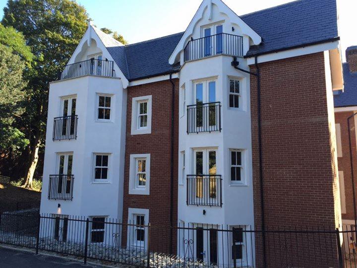 Elewacja nowo wybudowanego budynku mieszkalnego z balkonami i czarnymi balustradami, z fragmentem ogrodzenia w tle.