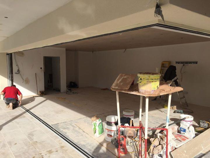 Mężczyzna w czerwonej koszulce instaluje prowadnicę do przesuwnych drzwi tarasowych na jasnej, kafelkowej podłodze; w tle surowy stan wykończenia wnętrza z widoczną instalacją elektryczną...