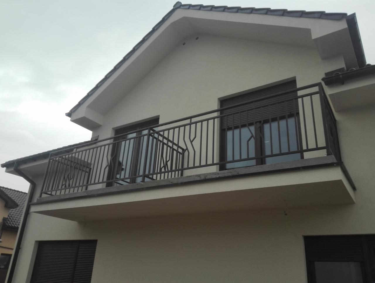 Czarna, metalowa balustrada balkonowa z dekoracyjnym motywem, zamontowana na balkonie budynku z beżową elewacją i brązowymi roletami.