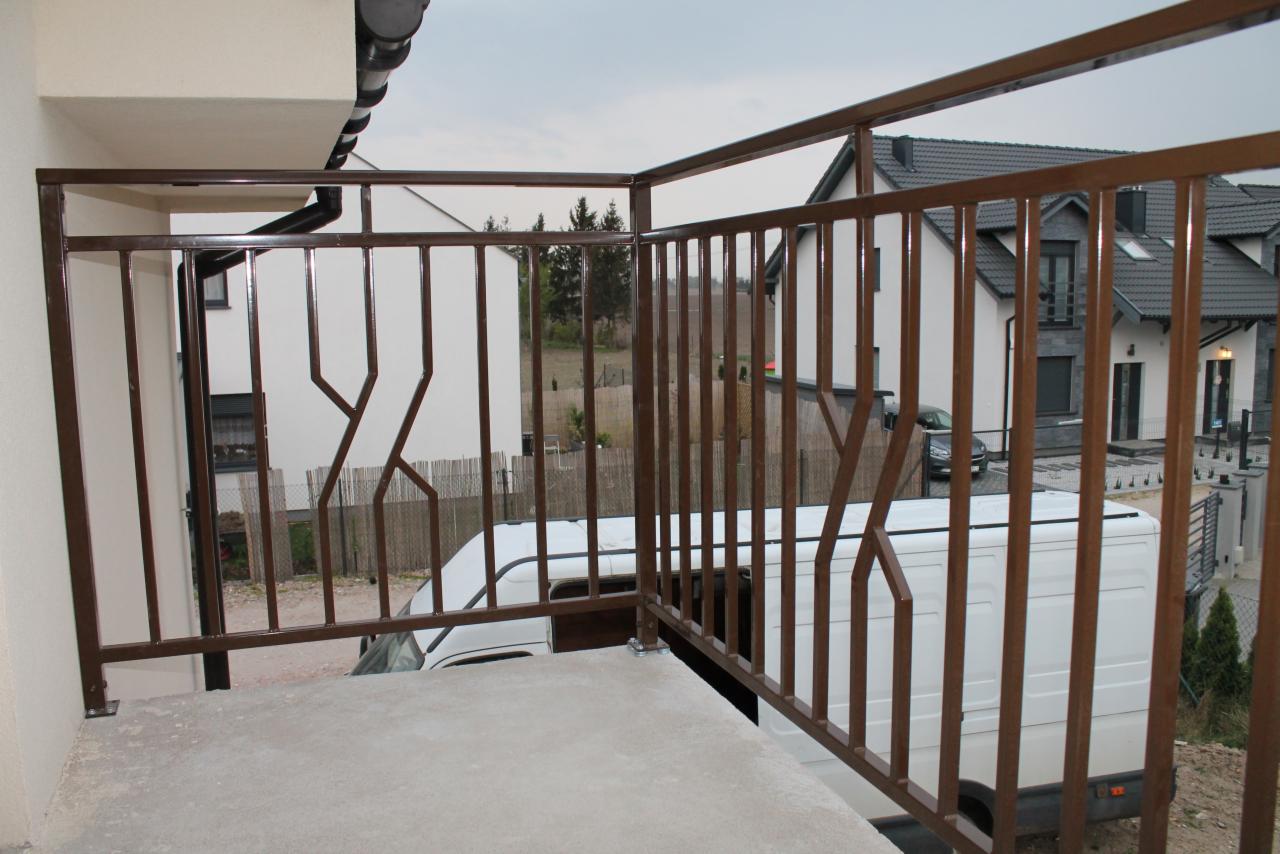 Balkon z brązową metalową barierką o geometrycznym wzorze na tle białego vana i domów.