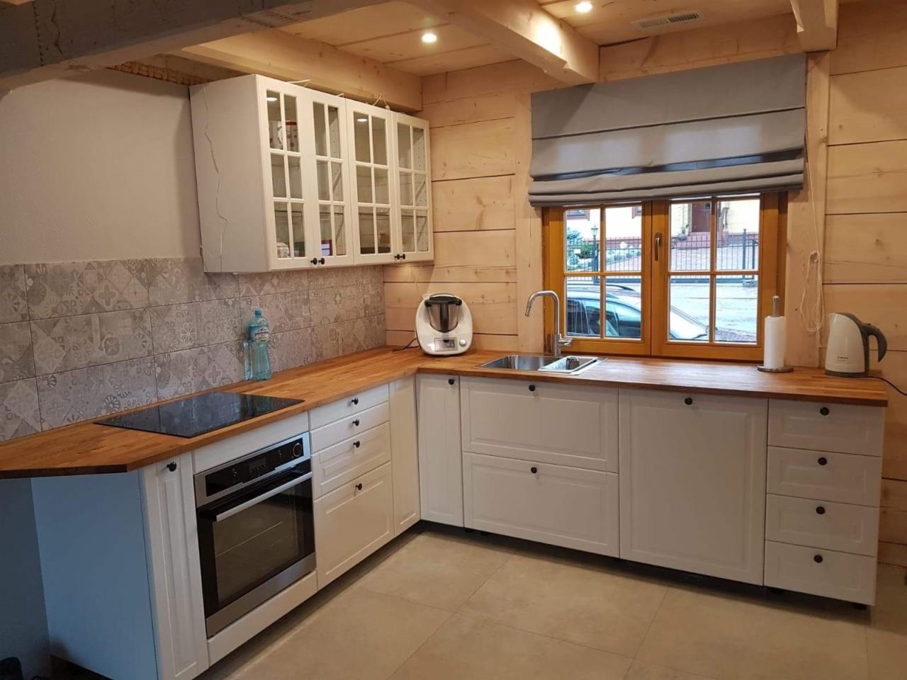 Kuchenna zabudowa w rustykalnym stylu: białe szafki z drewnianym blatem, okno z szarą roletą rzymską i drewniane ściany z widocznymi belkami sufitowymi, oświetlenie podszafkowe.