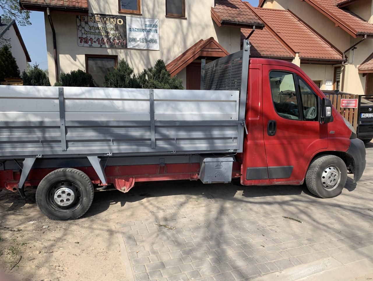 Czerwony samochód dostawczy z otwartą skrzynią ładunkową, zaparkowany na kostce brukowej przed budynkiem z szyldem reklamującym usługi związane z drewnem opałowym i biurem rachunkowym.