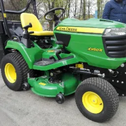 Zielony traktor ogrodowy John Deere X950R z żółtym siedzeniem i czarnym koszem na trawę, stojący na betonowej powierzchni, z widoczną osobą w tle.