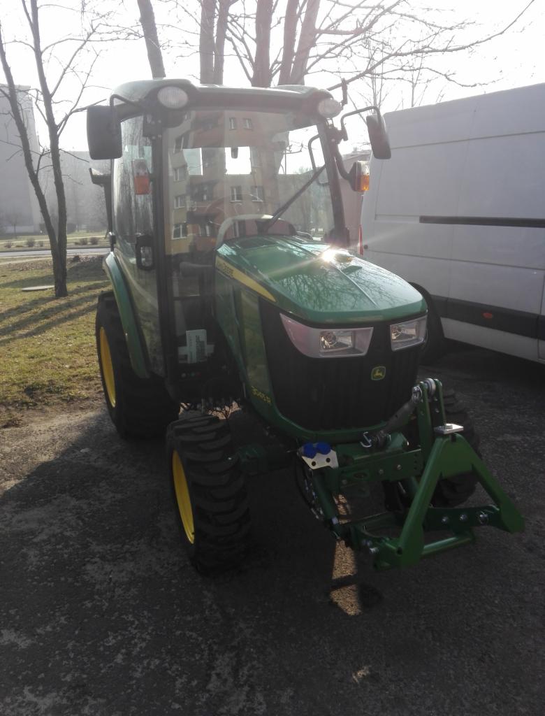 Zielony traktor John Deere 3005R z kabiną w Katowicach, stojący na żwirowym podłożu, z budynkiem w tle i częściowo widocznym białym busem po prawej stronie.