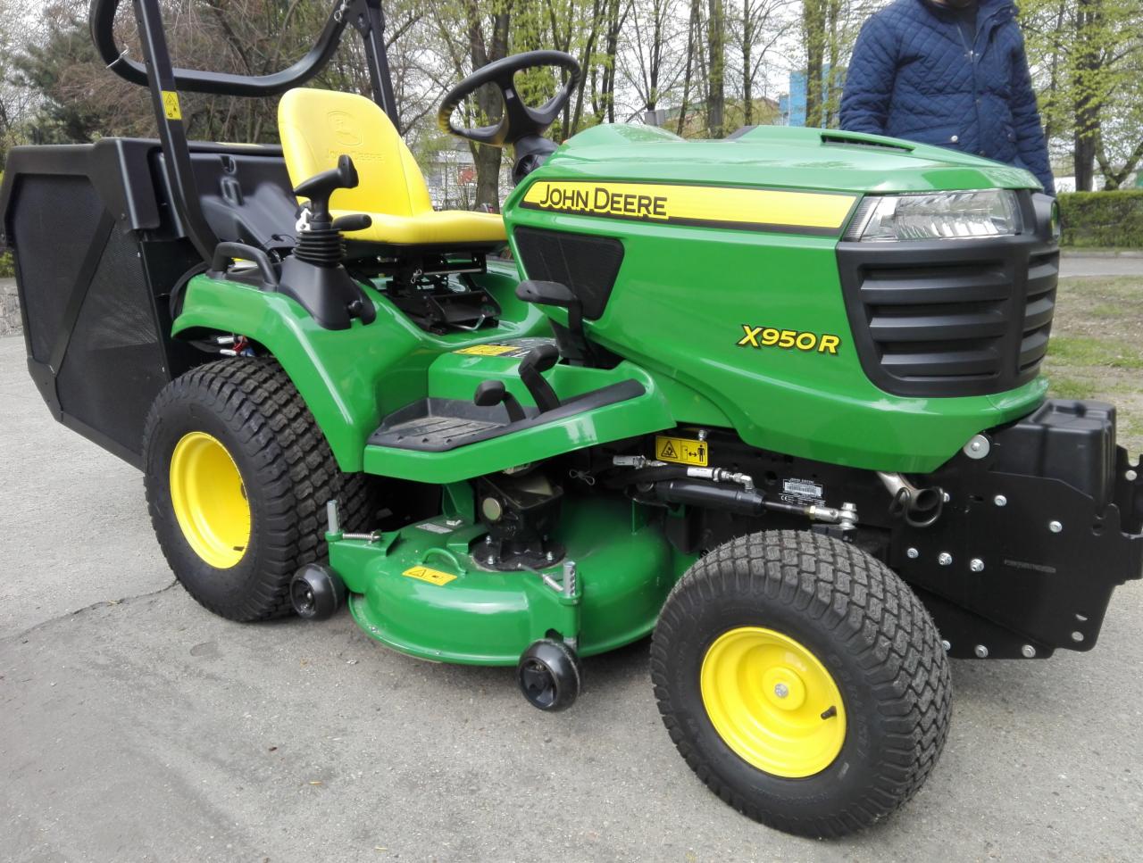 Zielony traktor ogrodowy John Deere X950R z żółtym siedzeniem i czarnym koszem na trawę, stojący na betonowej powierzchni, z widoczną osobą w tle.