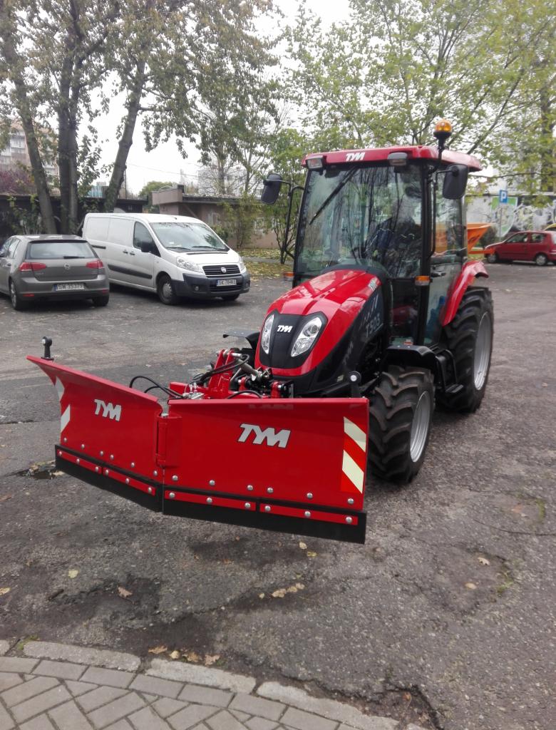 Czerwony traktor marki TYM z zamontowanym pługiem śnieżnym na tle miejskiego parkingu z drzewami i samochodami.
